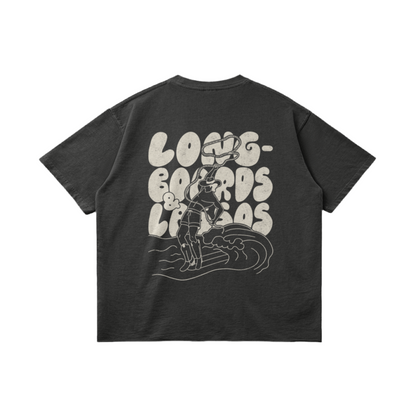 Back of a dark grey t-shirt with a white picture of a male surfer wearing cowboy boots and holding a lasso riding a wave on a longboard with the bubble writing saying, 'Longboards & Lassos' in the background.